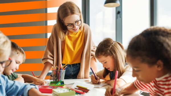 Professora observa atentamente um grupo de crianças da concentradas a desenhar e a pintar com lápis de cor numa sala de aula.