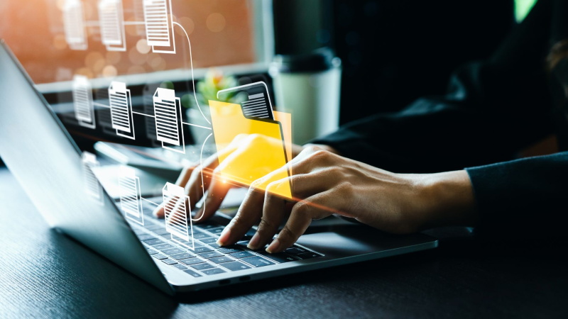 Pessoa a digitar num portátil com gráficos sobrepostos que simulam a organização e a transferência de ficheiros digitais para uma pasta amarela flutuante.